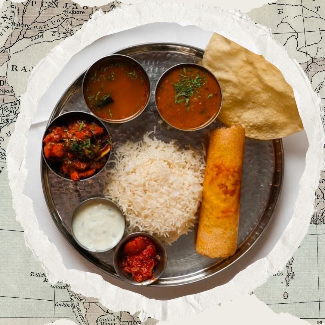A steel lunch thali seen from above: papadum, dosa, rice, curries, raita and pickle.