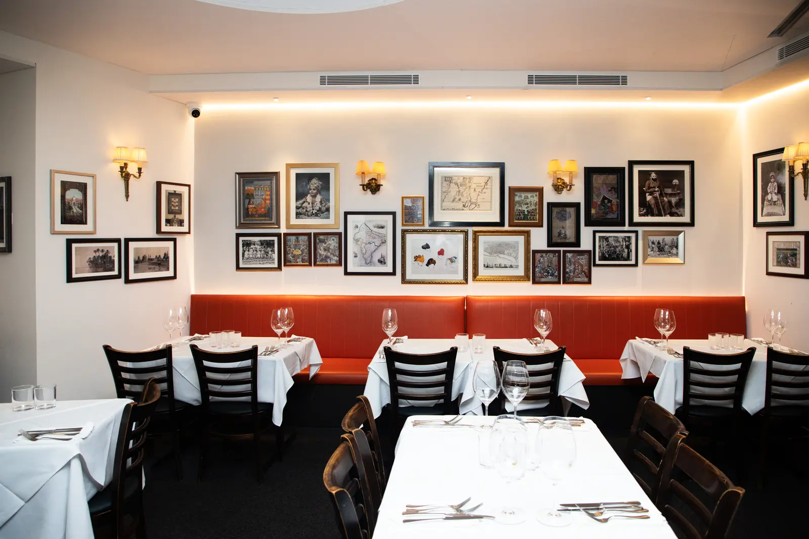 A long table laid with wine glasses and cutlery in front of an orange banquette and a wall of framed prints.