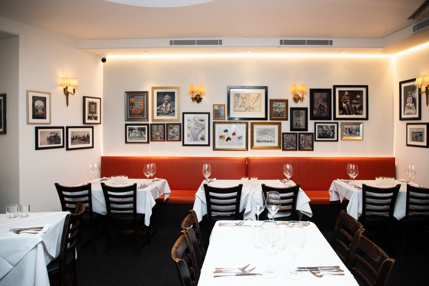 The Malabar dining room set with white tablecloths and wine glasses, in front of a red banquette and a wall of framed prints.