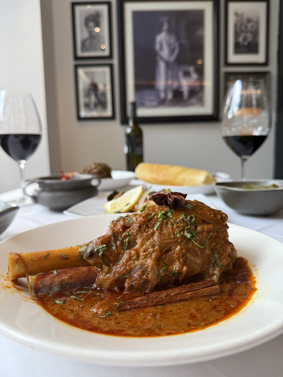 Lamb Shank Roganjosh on a white plate at a Malabar table, with glasses of red wine and framed prints behind.